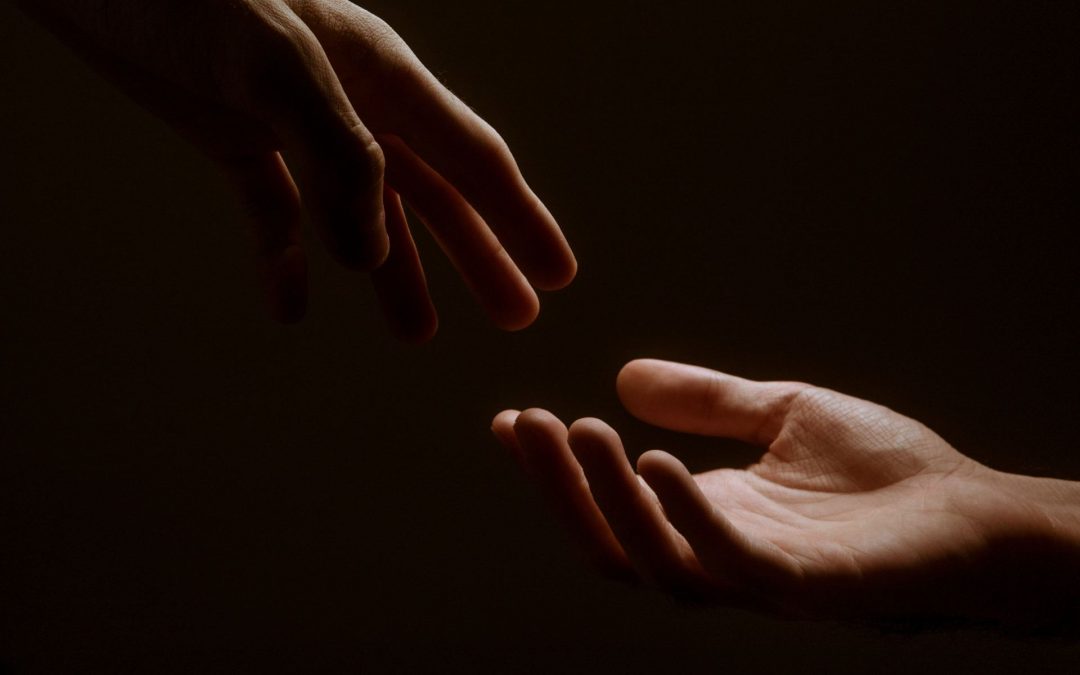 Two hands reach towards one another almost touching, against a shadowy black background